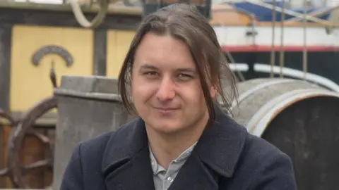 Jamie Niblock/BBC William Lund is looking towards the camera with a light smile. He has long hair in a ponytail, and is wearing a grey shirt and black coat. Behind him are boats docked.
