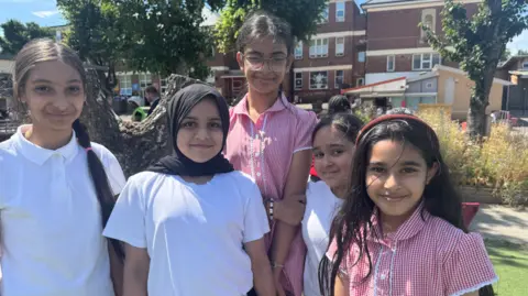 A group of five school girls stand looking at the camera. They are all smiling slightly. Two of them are in red gingham summer dresses. The others are wearing white t-shirts. In the background you can see trees, a playground and a school building.