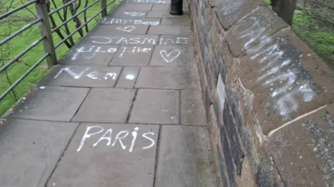 A stone walkway with a low wall on one side and a metal railing on the other. On the wall are words in white paint including 'Jasmine', 'Lilly' and 'Paris'. 