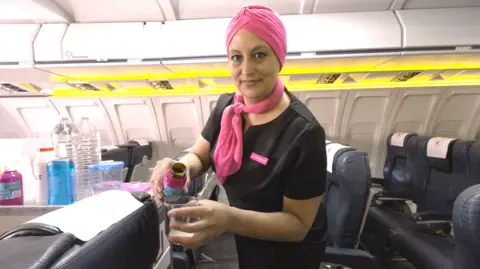 Chloe Jacquet Chloe Jacquet is dressed as an airline cabin crew, with a black dress, pink scarf and pink turban. She is leaning over a passenger's seat as she pours a drink into a glass.