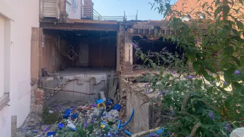 A disused building site with debris on the ground and overgrown weeds. 
