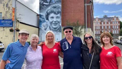 A group of people standing together in front of a mural of a women on it. Surrounding them are lots of buildings. 