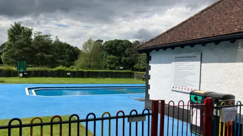 A small paddling pool surrounded by blue paving and a fence. There is a building next to it with pool rules written on the side. 
