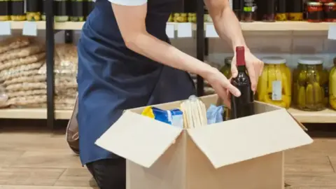 A man in blue overalls is kneeling in front of a shelf full of food. He is filling in a box with food and drink items.