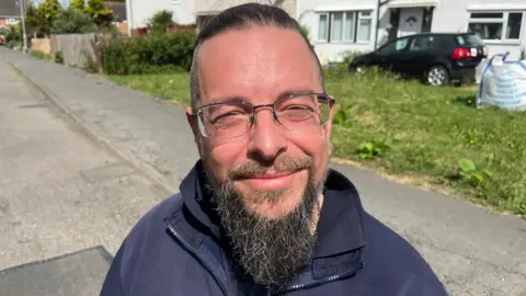 Mark Robinson is outside on the road in a residential area. He has glasses and a large beard. He has short brown hair and is wearing a blue fleece. 