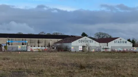 BBC/ Libby Merrywood The exterior of Brandon Stadium. The main public entrance is on the right of the image. Its gates are locked and boarded over, with hazard warning signs fixed to it. on the right of the image are two hangar-style structures, one with 'Coventry' painted on it, the other with 'Stadium' on it in blue paint.