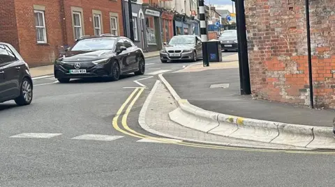 A view of the kerb on a corner with vehicles passing by next to it. On the other side of the road there are shops.