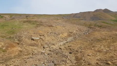 Environment Agency Ground covered in soil and rocks, partially covered by grass. Hills can be seen in the background.