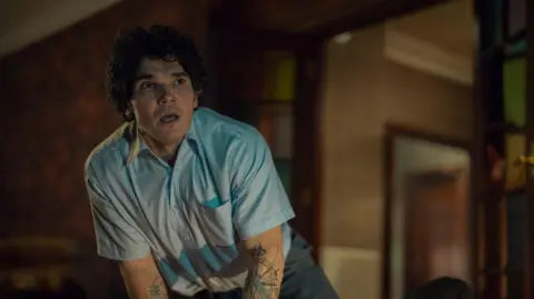 Netflix Fra Fee, wearing a short sleeve blue shirt. He has short, curly brown hair. 
