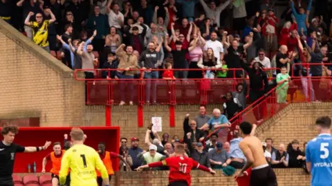Derek Lloyd A Droylsden player, in red kit, runs towards a stand with his arms outstretched in celebration as the goalkeeper, in yellow kit, jogs behind