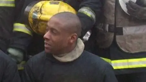 Jez Daniels Jez Daniels in his firefighter's uniform, holding a yellow helmet and with colleagues behind him