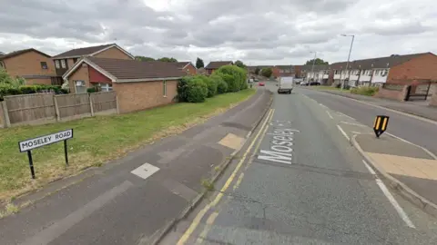 A picture of road  with houses on either side. There is also a pedestrian island with tactile paving and a sign with the road name. There is a white van and red car on the road in the distance. 