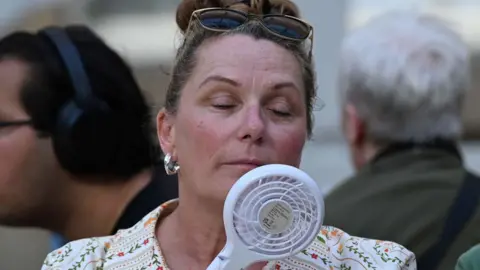 A woman cools her face with a hand-held electric fan