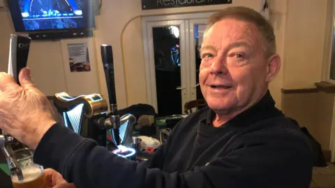 Kevin McCourt pulling a pint behind the bar in Cassanos nightclub. He is wearing a dark fleece and dark shirt, he has short sandy coloured hair and a TV screen can be seen on the wall behind him. He is looking to the right of the camera. 