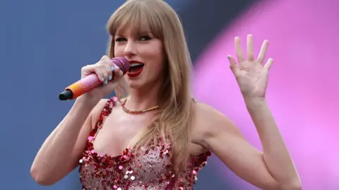 PA Media Taylor Swift holds her splayed hand out to the side of her, while clasping an orange and pink diamante microphone in the other. She is wearing a diamante and sequinned pink and silver corset and a gold necklace.