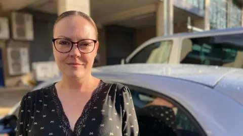 A woman wearing glasses looking down the camera lens. A car can be seen in the background. 