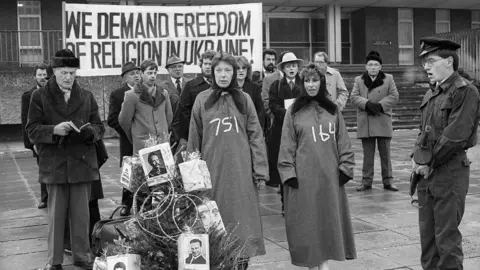 Tim Smith A black and white photograph of two women dressed in black headscarves and sackcloth robes with white numbers stencilled on then as two men dressed as guards look on.
