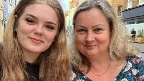 Two women smile at the camera. On the left a teenage girl with blonde hair and on the right a middle aged lady with a floral top smiles at the camera. She also has blonde hair.