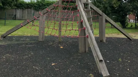 Friends of Portswood Rec Two children perch on the top of a wooden climbing frame in a park. On one side of the climbing frame there is a large rope ladder. In the background there is grass and trees.