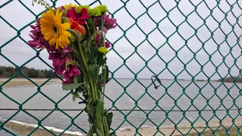 Henry Godfrey-Evans/BBC A floral bouquet that has been strapped to a metal fence. It is made of flowers that are different shades of yellow, orange, pink and red. A reservoir can be seen on the other side of the fence. It is a grey cloudy day.