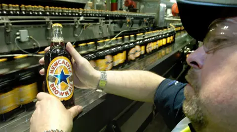 A worker at the Federation Brewery looks at a bottle of Newcastle Brown Ale as hundreds of other bottles whizz past on the production line. The bottle is made of glass and contains a dark liquid. The label says "Newcastle Brown Ale" and has a blue star in the centre.