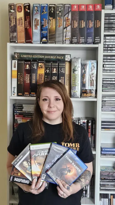 Amy Dyson A woman wearing a Tomb Branded t-shirt is stood in front of a tall set of shelves which are stacked with Tomb Raider games.