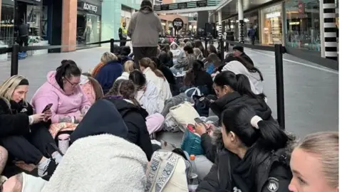 A long line of people sit in between barriers in a queue before the store opened