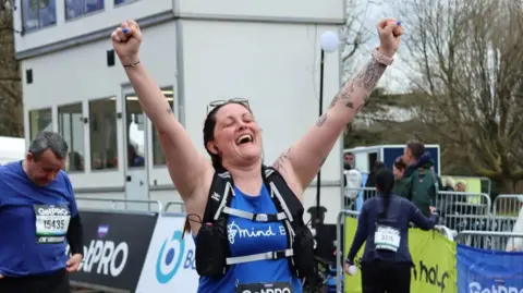 Alicia wearing a running bib and Bath Mind branded tank top with her hands in the air at the Bath Half Marathon finish line.
