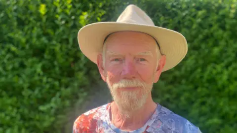 BBC / Elizabeth Baines A man with a white beard and straw hat smiles at the camera. He wears a blue top and behind him green space is blurred.