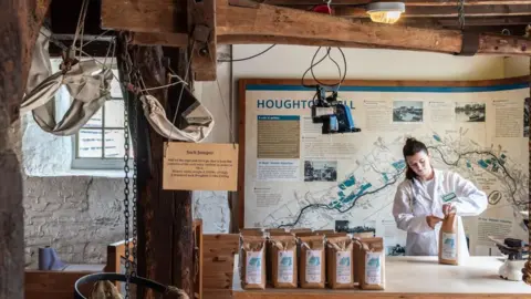 A woman in a white coat puts flour into brown paper bags inside Houghton Mill.