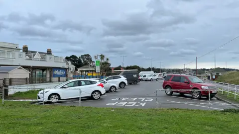 BBC A small seaside car park with several parked vehicles, including white and red cars, bordered by metal railings. The foreground shows a grassy area and the background features low-rise buildings with signage, a cloudy sky, and distant streetlights.