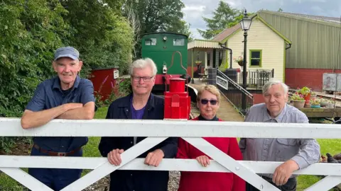 Volunteers stand at a gate in front of a train and station house on the East Wressle and Brind Railway