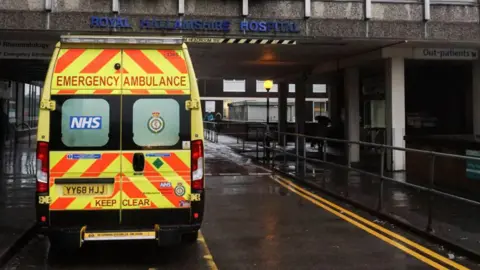 Getty Images An NHS emergency ambulance is parked outside the entrance of Royal Hallamshire Hospital on a wet roadway. The rear of the ambulance displays high-visibility yellow and red markings, along with the text “EMERGENCY AMBULANCE” and “KEEP CLEAR.” Hospital signage, covered walkways, and railings are visible in the background, indicating a medical facility entrance area.