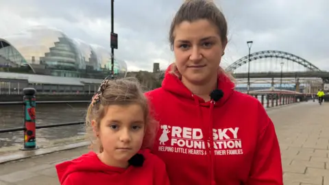 BBC/Jim Scott Beth Graham, who is wearing a red hoodie with white Red Sky Foundation lettering, along with her daughter, Heidi, who is also wearing the same hoodie, are on the Newcastle Quayside. Behind them can be seen the Tyne Bridge to one side and The Glasshouse International Centre for Music to the other, on the Gateshead side of the River Tyne.