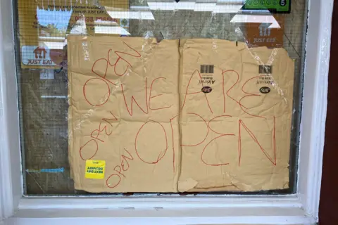 A piece of cardboard with red writing on it saying 'We are open' and several uses of 'open' on it. The cardboard is stuck up in a broken window pane.