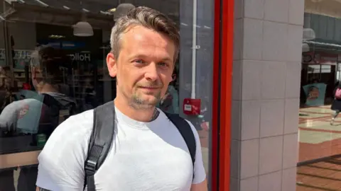 A man in a white t-shirt with a backpack on his back. He is standing in front of a shop. 
