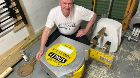 John Devine/BBC Steve Wainwright is in his garage with a large tape measure and plug next to him.
