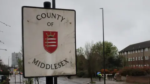 LDRS A white street sign reading COUNTY OF MIDDLESEX with a red coat of arms in the centre. The sign is beside a main road