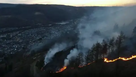 A line of flames can be seen going across a hill overlooking Treorchy town down in the valley below. Heavy smoke can be seen rising into the air, with trees visible in front, and protruding through the gloom. It is dusk, and a sunset can be seen in the horizon. 