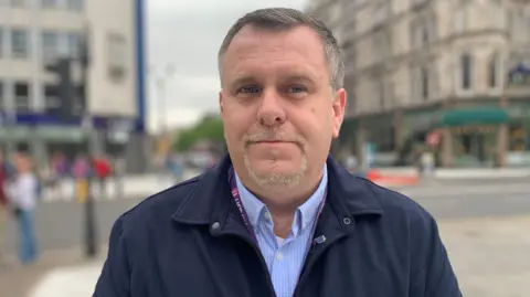 Liam English with short grey hair and a goatee beard looks at the camera. He is wearing a navy coat with a light blue striped shirt underneath.