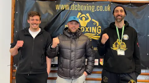 David, Alex, and Ian from DC Boxing are stood in front of one of their banners. Each is posing with their fist up and smiling.