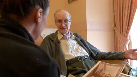 Ron Champion is in the background sitting in his armchair. He's wearing a pale shirt, grey cardigan and grey trousers. In the foreground his befriender, Sarah, has her back turned towards Ron and a sepia photograph in her hand.
