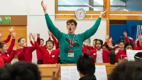 Children rehearsing songs with tutor. They have their hands in the air. The children are wearing red uniforms. A man is seen playing the piano. There is a clock on the wall behind them.