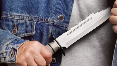 A youngster holds a large serrated blade across his chest. He has one a grey T'shirt and denim jacket.