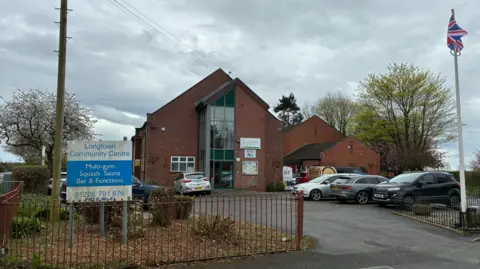 An exterior view of Longtown Community Centre. The building is a large, red brick structure with a long vertical window, iron railings and a sign outside with the centre name on it. 