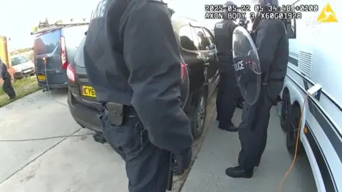 Warwickshire Police A still from body camera footage that shows police officers dressed all in black, standing between a black car and a white caravan. One officer is holding a shield and an orange cable is trailing from the side of the caravan. A grey van is to the left of the black car.