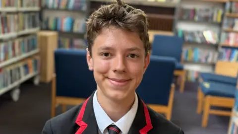 Qays Najm/BBC Daniel sitting in his school library. He has short brown hair, is wearing his black and red school uniform and is smiling.