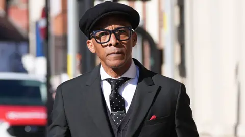 Image shows Jay Blades arriving at Worcester Crown Court wearing a black suit, white shirt, black tie with white spots and a tie pin and a red handkerchief tucked into his breast pocket. He is wearing a black peaked cap.