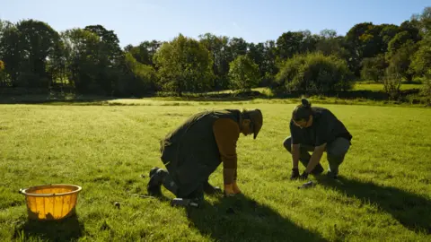 LDNPA Two people crouched on some grass who look to be planting. There is a yellow bucket beside them. It is a sunny day with a clear blue sky and the grass looks very bright. There is a line of trees in the background.