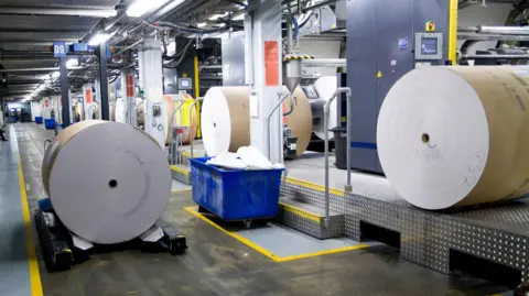 Getty Images A giant roll of white paper (newsprint) sits on a wheeled transport cradle in the centre-left foreground. A blue, wheeled container contains discarded sheets of white paper.
